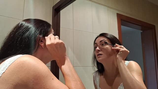 Woman applies mascara while looking in a bathroom mirror before getting ready