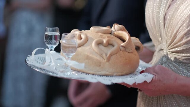 A woman holding a tray with a traditional decorated bread loaf and two shot glasses at a wedding.