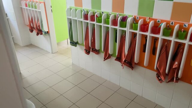 School restroom or washroom corner featuring row of white wall-mounted sinks at left and repeating cubby hooks with orange fabric bags on right.
