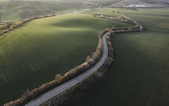Aerial view of the winding Vlnky road cutting through lush green rolling fields in Nasedlovice, South Moravian Region, Czechia.