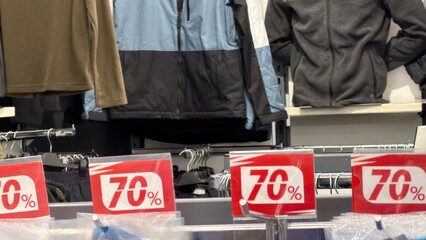 Close-up of red “70%” sale signs on a clothing rack  jackets and fleece tops are visible in the soft‑blurred background. Retail store interior, artificial lighting, shallow depth of field emphasizing  © valentina fridman