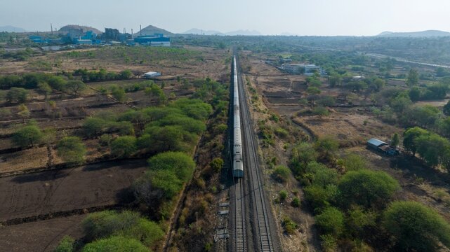 Aerial view of a passenger train traveling along railway tracks near the Agra - Mumbai Road, surrounded by rural fields and industrial buildings, Nashik, Maharashtra, India.
