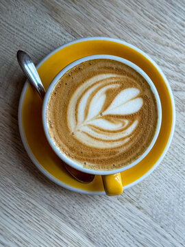 Cappuccino with Latte Art Rosetta in Yellow Ceramic Cup on Wooden Table. A high-detail vertical top-down photograph of a freshly brewed cappuccino or flat white featuring a precise rosetta leaf latte 