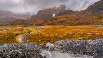 Autumn mountain valley with waterfalls and rocky stream in Norway  A rocky stream cascades over the foreground into an autumn mountain valley in Norway. A distant waterfall descends from rugged peaks  © olekristian