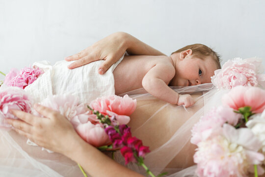 Cute newborn baby lying on mother belly with peony flowers. Love and care concept. Portrait of baby son on mom tummy, tender moment. Adoption and maternity. Happy Mothers day.