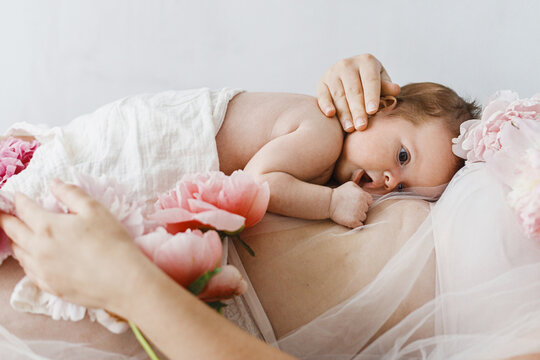 Cute newborn baby lying on mother belly with peony flowers. Love and care concept. Portrait of baby son on mom tummy, tender moment. Adoption and maternity. Happy Mothers day.