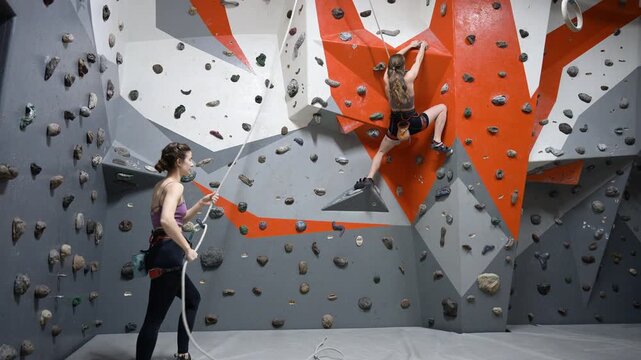 Female athlete climbs boulder route in gym with supportive friend standing below on mat. Dynamic stock video for strength, coordination, determination, and teamwork concepts