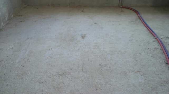 Camera pulls back to reveal an empty room during renovation. Unfinished concrete walls, floor screed, new white radiator and red and blue water pipes for the heating system. Nobody