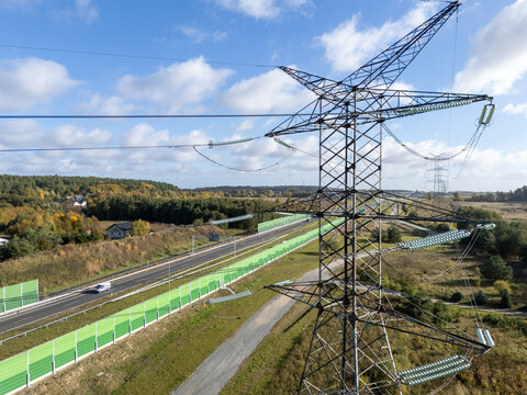 Aerial view of a high-voltage electricity pylon standing beside a highway with green noise barriers, surrounded by autumn forest and fields in Gdansk, Poland.