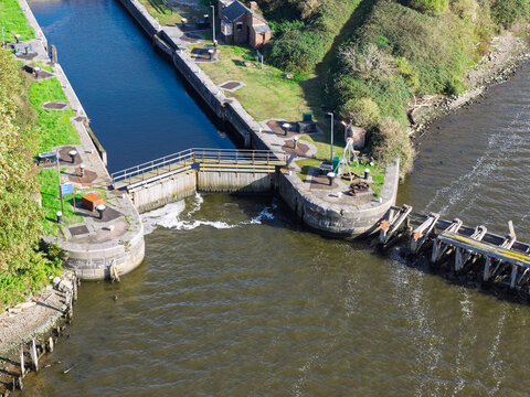 Aerial view of a canal lock system with stone and concrete walls, wooden gates, and calm water flowing through the channel in the United Kingdom.