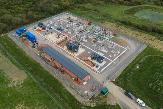 Aerial view of an electrical substation featuring transformers, switchgear, and maintenance buildings situated within a fenced compound surrounded by green fields in the United Kingdom.