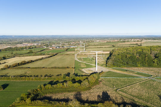 Aerial view of a T-shaped electricity pylon standing in a rural landscape with green fields, hedgerows, and trees in the United Kingdom.
