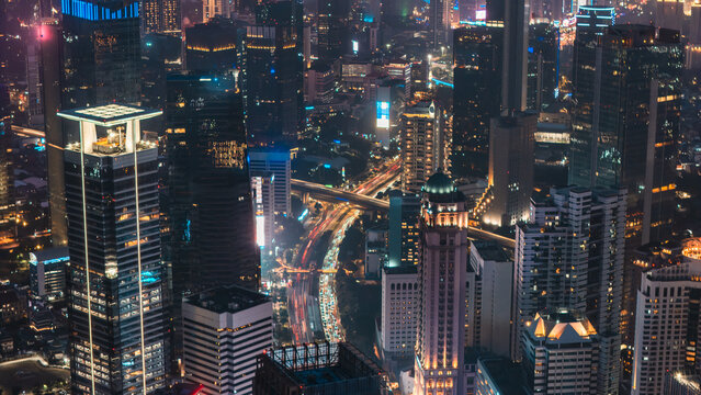 Aerial view of the illuminated Jakarta skyline at night with dense skyscrapers, glowing office buildings, and busy traffic trails on the roads below in Jakarta, Indonesia.