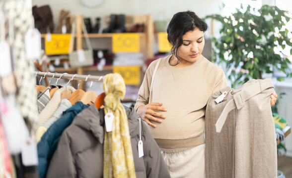 Pregnant Latina near the stand with warm clothes, she looks at beige pants for autumn and winter. Customer in the third trimester chooses comfortable trousers