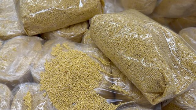 Close up of heaps of yellow millet groats packed in clear plastic bags on a store counter. Healthy grain cereal crop product for bulk culinary. Defective, damaged packaging and spilled grain.