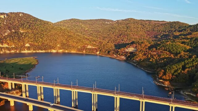A rail bridge crosses a lake bordered by forested hills in autumn, with the scene lit by the low sun in the late afternoon.