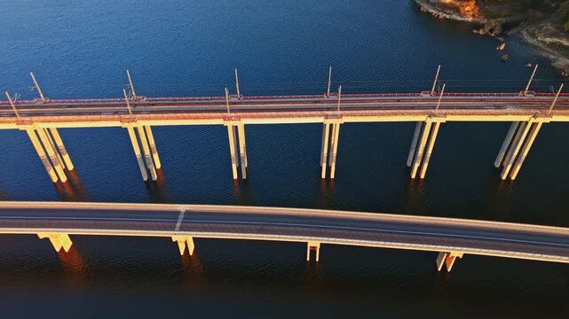 Two parallel bridges span blue water, with workers and equipment along the upper structure during daylight a small shoreline area is visible at the top right.