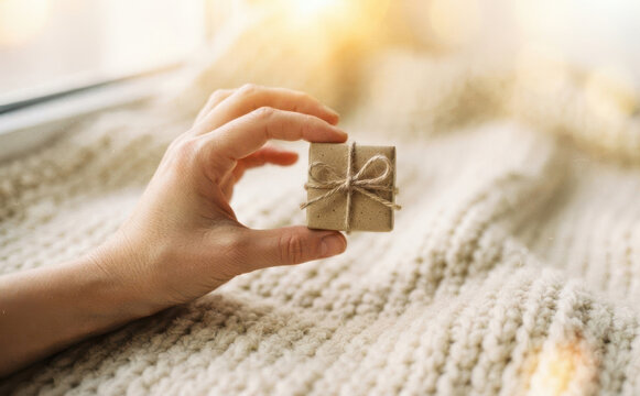 Woman holding a small gift box wrapped in craft paper with string bow over a cozy knitted blanket for holiday surprises