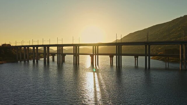 The sun sets behind a rail bridge crossing a river, casting a reflection on the water near a mountain slope at the end of the day.