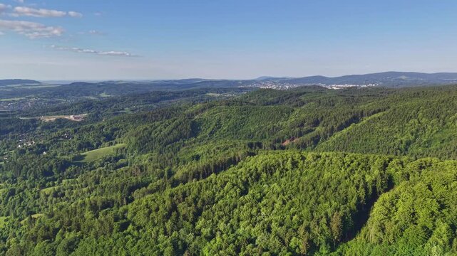 Sweeping aerial shot captures lush green hills blanketed by forest, gentle ridges, and sunlit valleys. Distant town sits on horizon under blue sky, creating tranquil natural landscape suitable