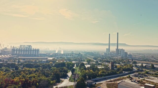 A power station with two smoke stacks stands beyond roads, trees, and buildings in a city landscape during daylight, with hills on the horizon.