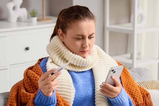 Telemedicine and healthcare access, sick woman checking body temperature with digital thermometer and smartphone at home during seasonal flu recovery
