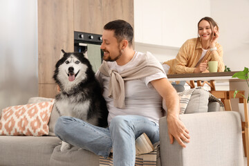 Pet ownership and domestic lifestyle, man sitting on sofa with husky dog while woman relaxes in background at home kitchen © Pixel-Shot
