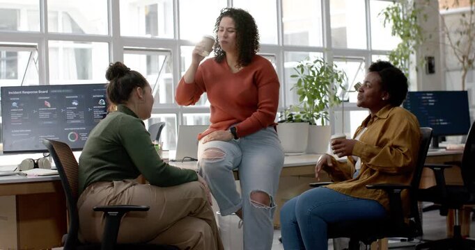 Female coworkers gathering at open office during coffee break, center woman in rust top sipping cup