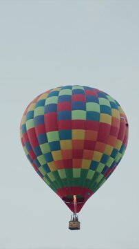Hot air balloon floats in sky with colorful checkered pattern and basket visible. Balloon rises steadily against overcast background flame burns at base of wickering basket