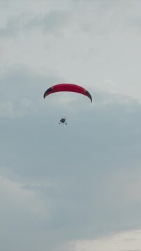 Red parachute and paratrike with visible frame glide through cloudy sky. Pilot operates under canopy equipment details like helmet seen. Sky dominates background with soft light clouds