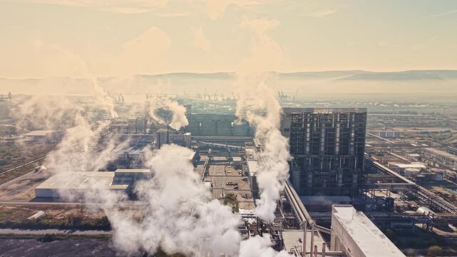 Steam rises from power plant stacks among factory buildings and roadways on the outskirts of a city, with the sun low in the sky.