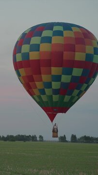 Hot air balloon floats above green landscape carrying passengers in its wickered fabric envelope. Basket hangs beneath colorfulful checkered balloon against sky backdrop