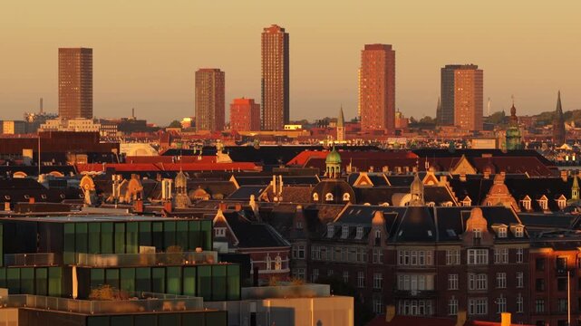 Aerial drone view of high-rise towers on the horizon above old town rooftops and green copper domes in golden light, Copenhagen, Denmark.