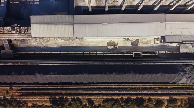 Railway tracks run beside a long covered platform and a freight train in an overhead view of a station during daytime.