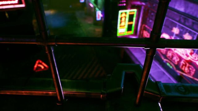 Steel metal pipes form a safety barrier above a dark platform lit by bright red and green neon signs during the night hours in a city area