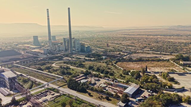 Aerial view of a power plant and industrial complex with tall chimneys, pipelines, and roads near a city in daylight, with a flat landscape extending to the horizon.