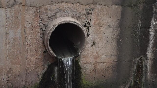 Water pours from a round concrete drainage pipe embedded in a cracked wall, with dark moisture stains and green growth beneath the outlet.