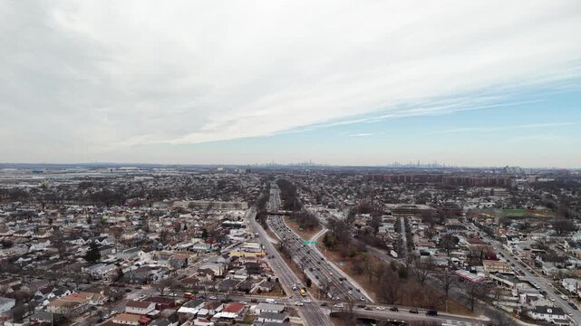 A wide drone pan crosses a divided highway and winter neighborhoods in Queens. Multiple traffic lanes curve through a dense pattern of roofs and leafless trees beneath a pale sky with thin clouds.