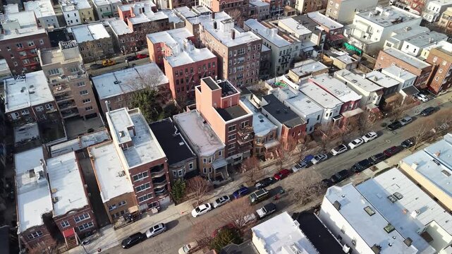 An angled drone glide crosses brick houses and a tree lined street in Queens. Flat roofs and apartment facades fill the view as the camera passes parked vehicles and bare trees in winter sunlight.