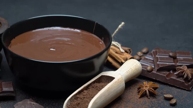 Melted chocolate swirling in a black bowl with cocoa powder, dark chocolate bars, cinnamon sticks, star anise, and coffee beans arranged on a dark surface