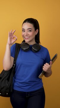 Happy female university student holding a tablet and making an ok gesture on a yellow background