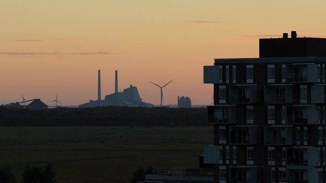 Aerial drone view of a residential tower silhouetted at sunset, the CopenHill power plant and wind turbines on the horizon, Copenhagen, Denmark.