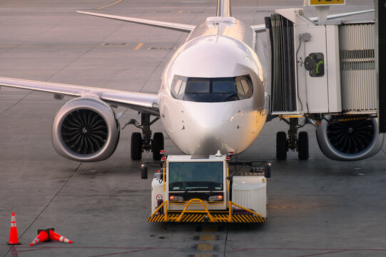 Front view of white commercial passenger airplane connected to jet bridge with pushback tractor on airport tarmac at golden hour