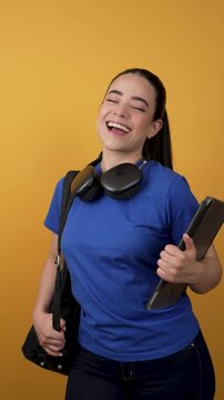 Young latina female student with headphones and backpack dancing joyfully against a yellow background