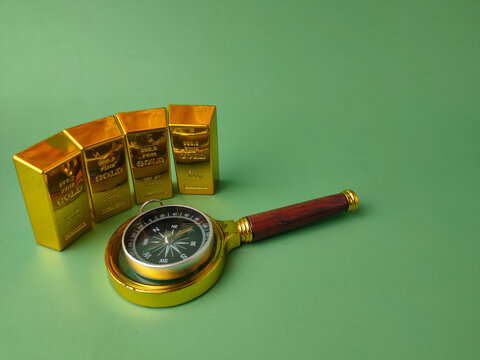 Four upright gold bullion bars stand behind a brass compass resting inside a magnifying glass on a green surface