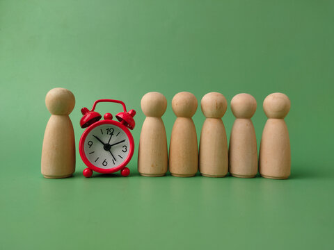 A small red alarm clock stands placed in a row of wooden peg dolls on a green backdrop