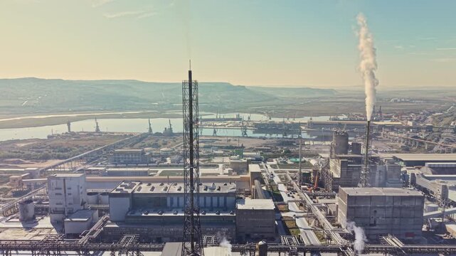 Buildings, pipes, and towers fill an industrial complex beside a body of water, with steam rising from a stack in daylight.