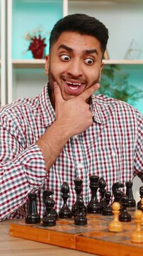 Indian man on home table plays chess and studies next move with calm smile and deep focus during board game. Arabian boy on home table thinks over strategy and enjoys calm leisure with brain training.