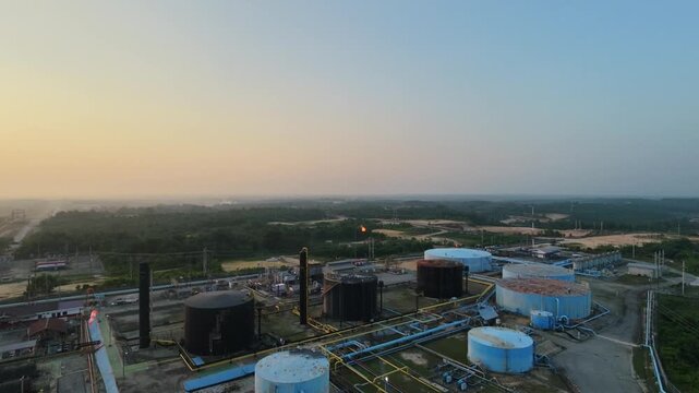 Aerial view of an industrial oil refinery plant with large storage tanks, piping systems, and processing facilities under a glowing sunset sky. Energy, fuel production, and industry concepts