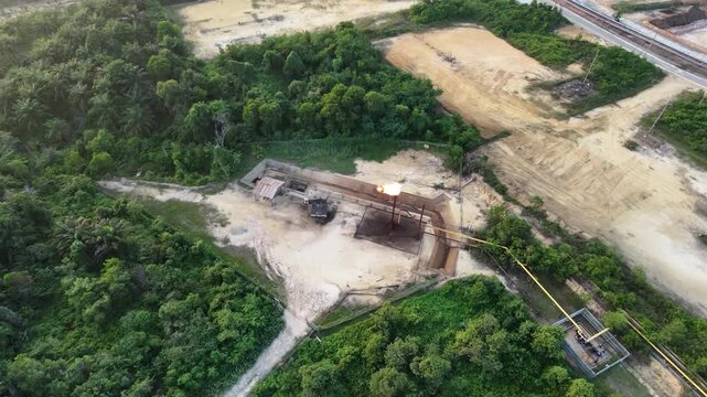 Aerial view of an industrial energy facility with processing units, pipelines, and a flare stack operating amidst lush green forest and open land. Industrial infrastructure and energy concept.